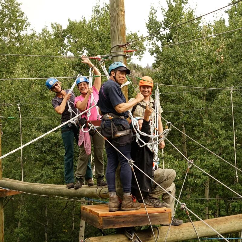 Four people are on a ropes course, suspended in the air. They are wearing helmets and safety harnesses, and appear to be navigating a wooden beam. The background features lush green trees, suggesting the course is set in a forest or park. The sky is overcast, providing soft, diffused lighting.